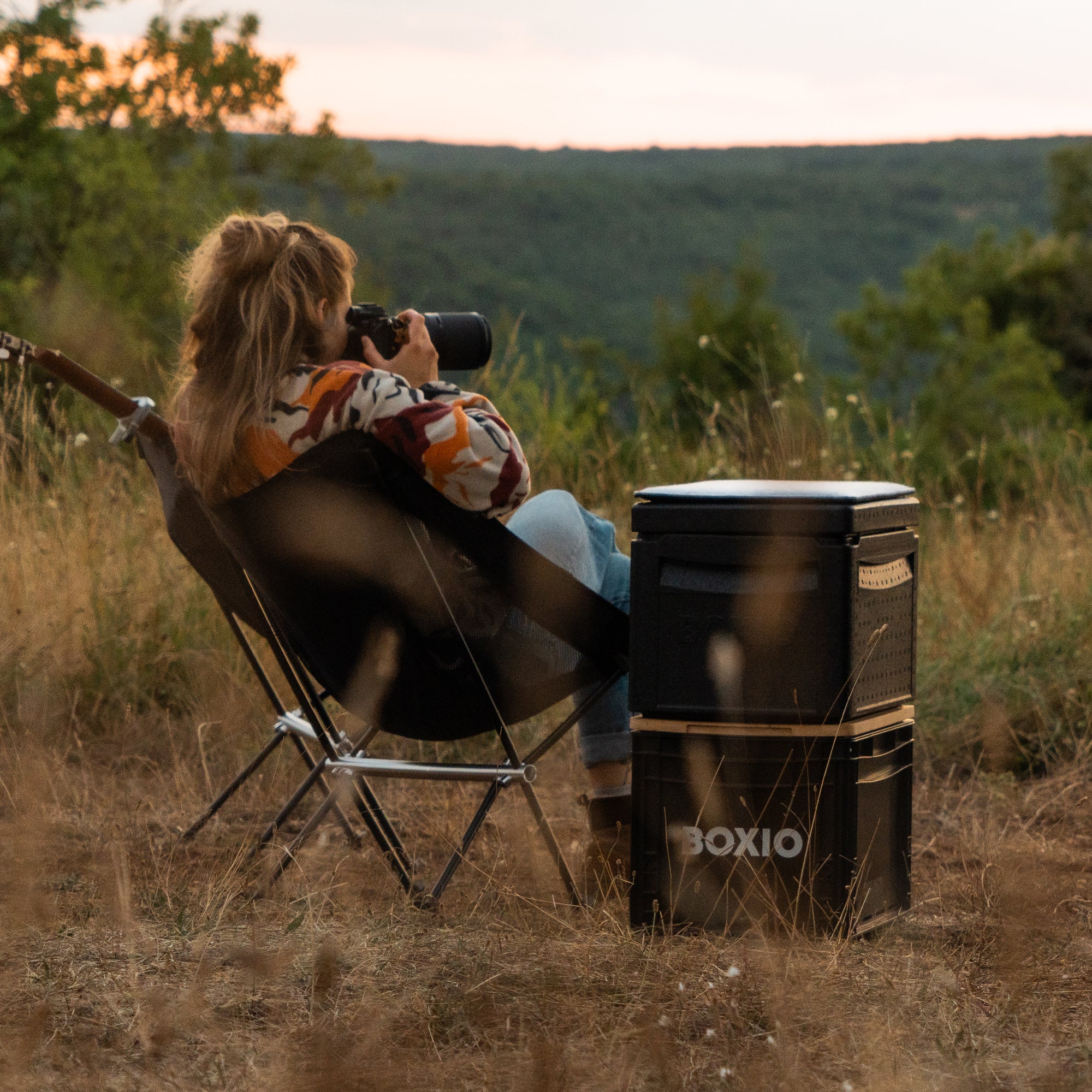 Frau mit Kamera sitzt in einem Campingstuhl im Freien, umgeben von Natur und einem Boxio-Kühler.