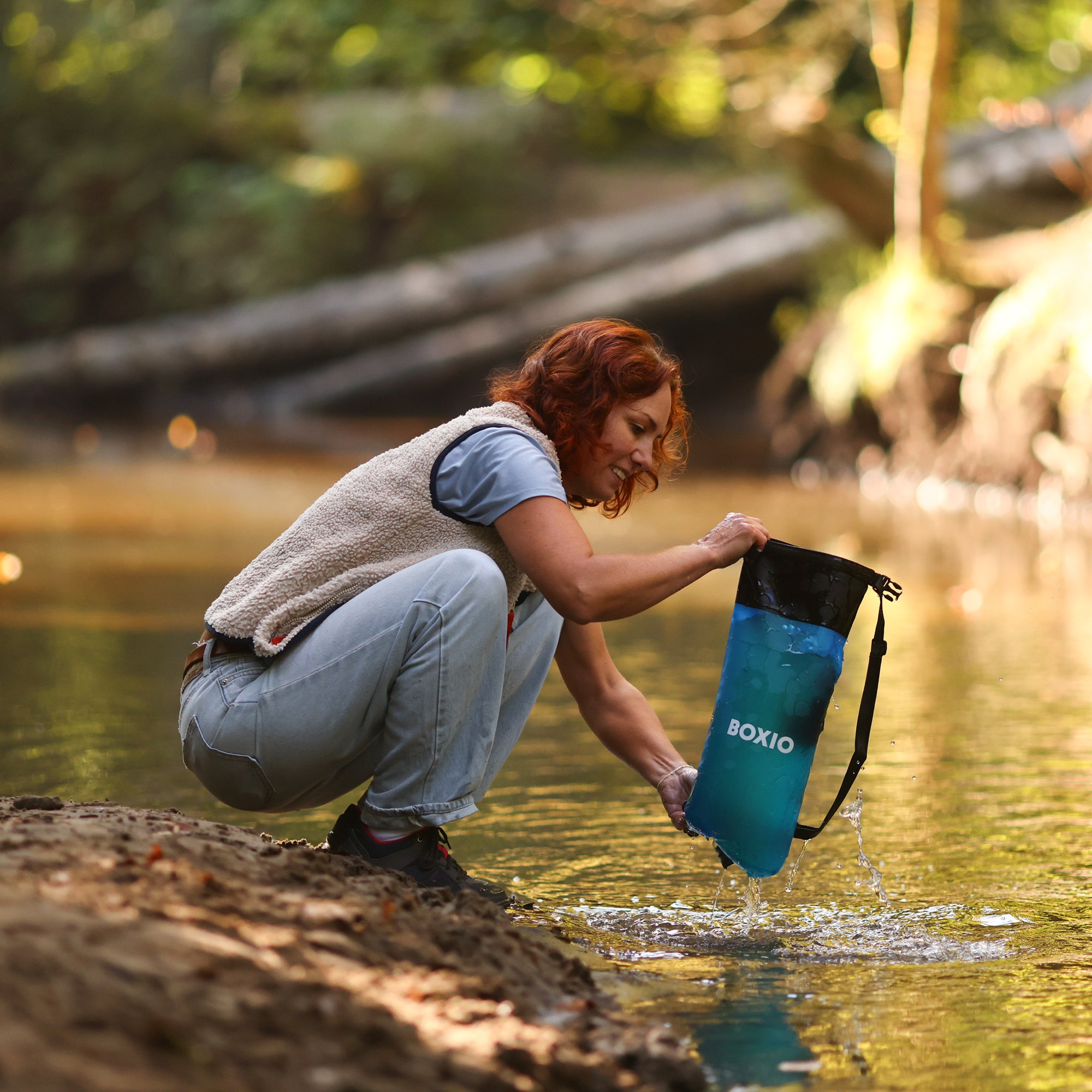 Frau mit roten Locken, die am Ufer eines Flusses Wasser in einen blauen Behälter schöpft.