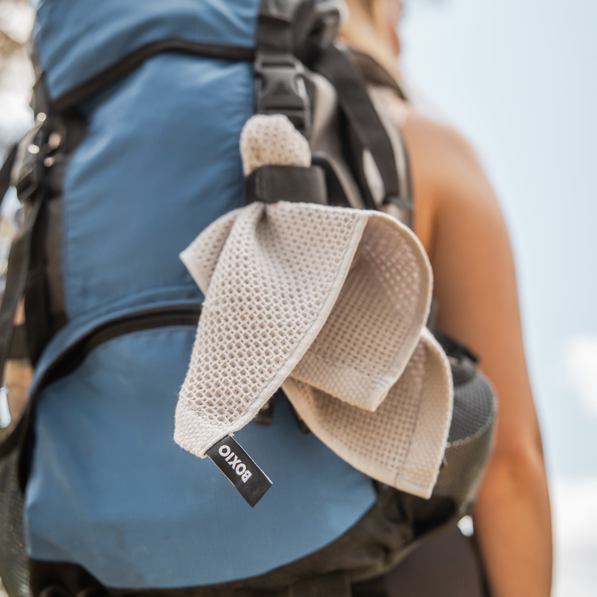 Grauer Sporttuch hängt an einem blauen Rucksack, während eine Wandererin im Hintergrund steht.