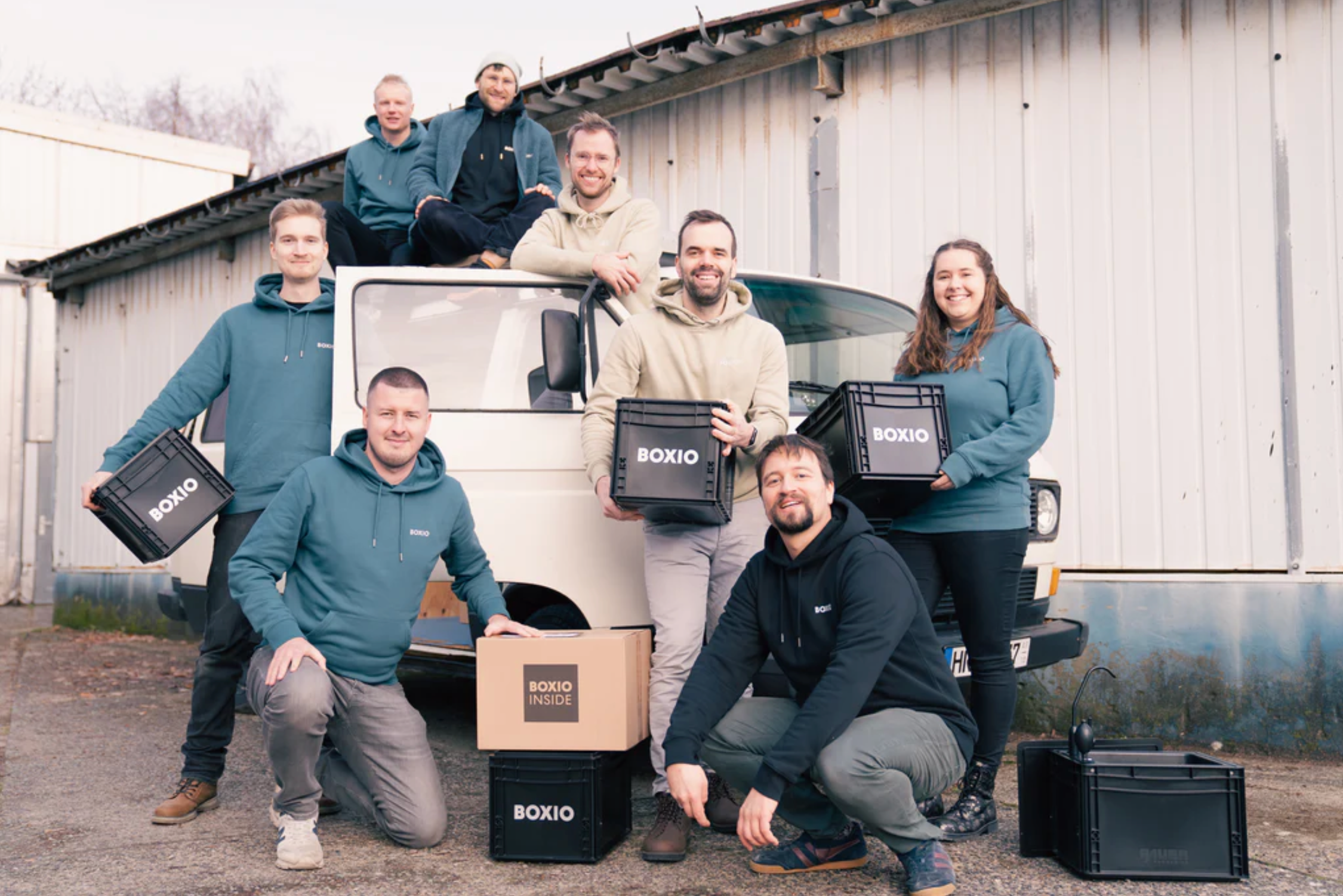 Gruppe von acht Personen in Sweatshirts mit der Aufschrift "BOXIO" vor einem Lieferwagen, einige halten Kartons und Kisten.