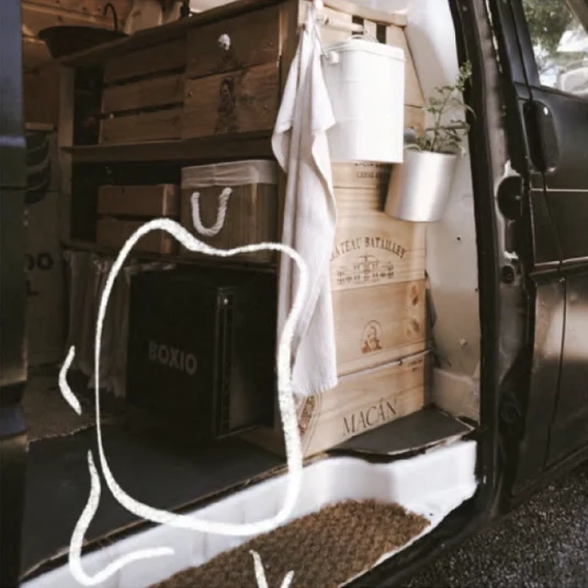 Innenansicht eines umgebauten Vans mit Regalen aus Holzkisten, einer Pflanze und einem schwarzen Kasten beschriftet mit "BOXIO".