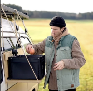 Mann mit Kappe und Weste legt einen Apfel in eine schwarze Box neben einem Van auf einem Feld.
