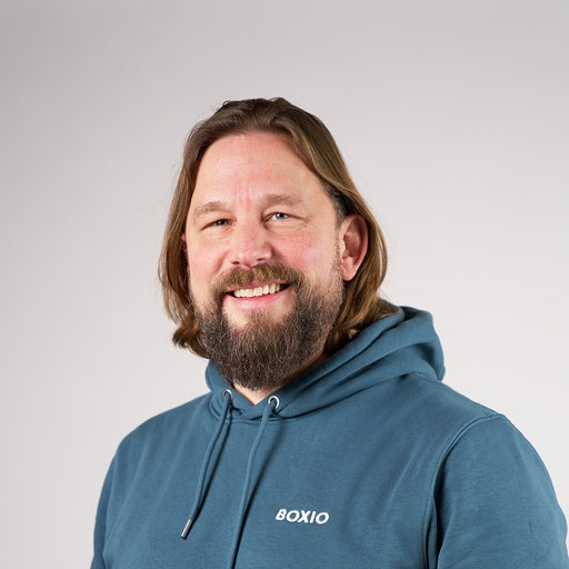 Mann mit schulterlangen Haaren und Bart trägt einen blauen Hoodie mit der Aufschrift "BOXIO" und lächelt freundlich.